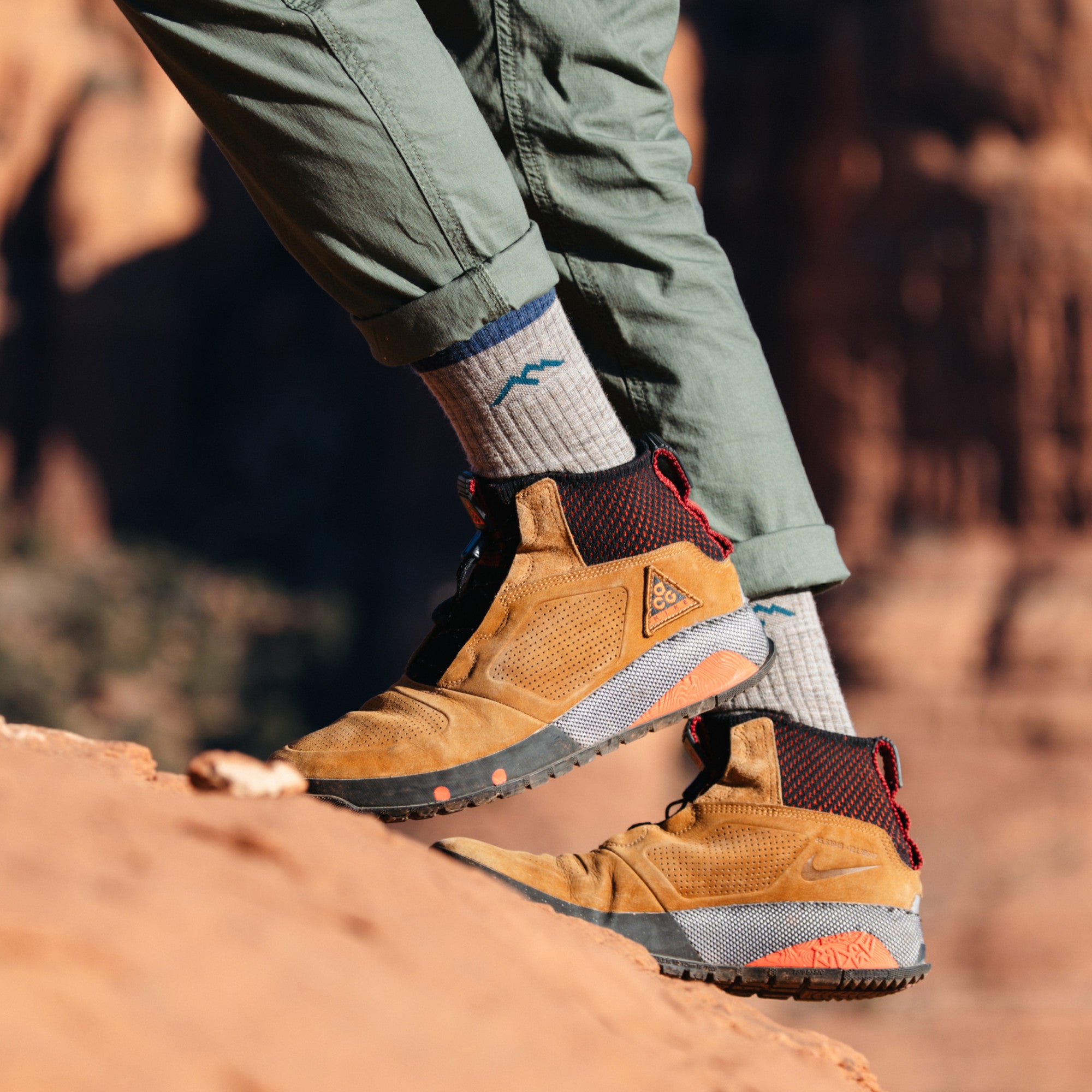 Close up shot of model wearing brown hiking boots and green pants in 1466 socks in rye colorway hiking on red desert rock