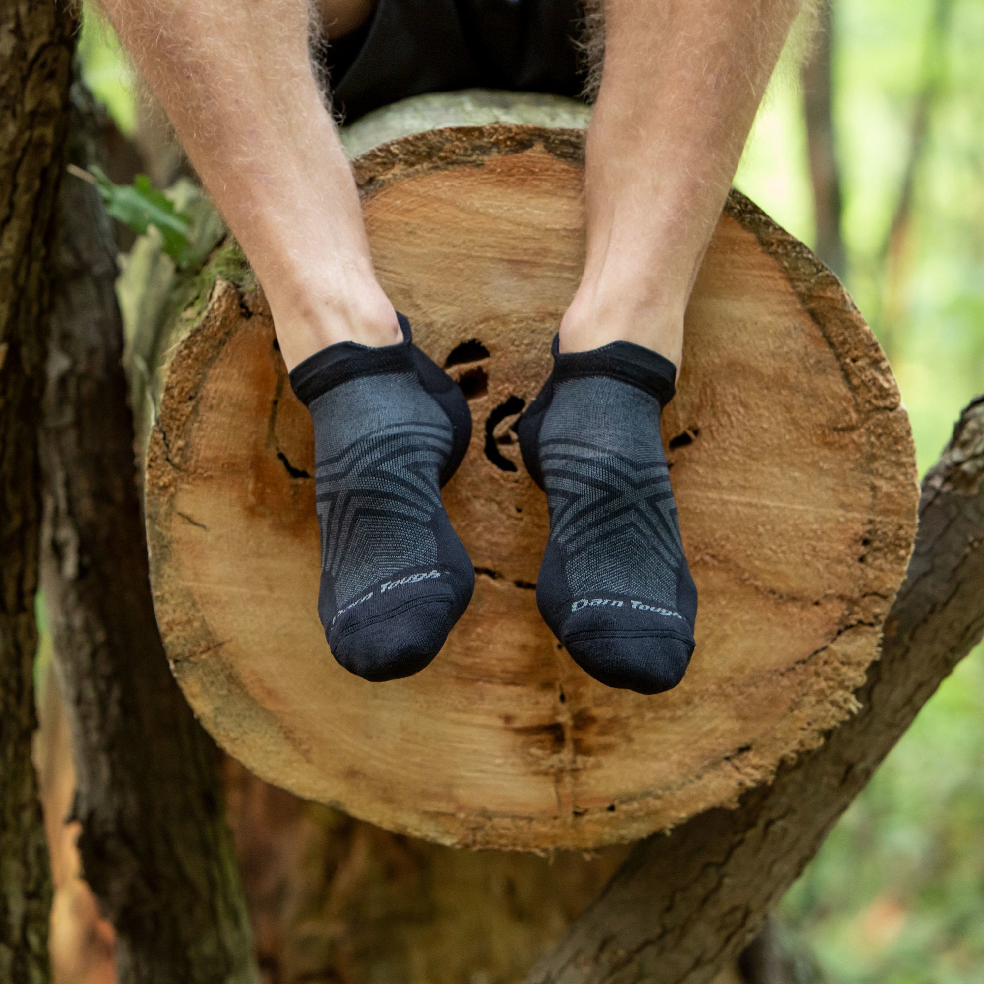Man sitting on a log in the woods, feet dangling, wearing Run No Show Tab Ultralightweight Running Socks in Black, Lifestyle Image