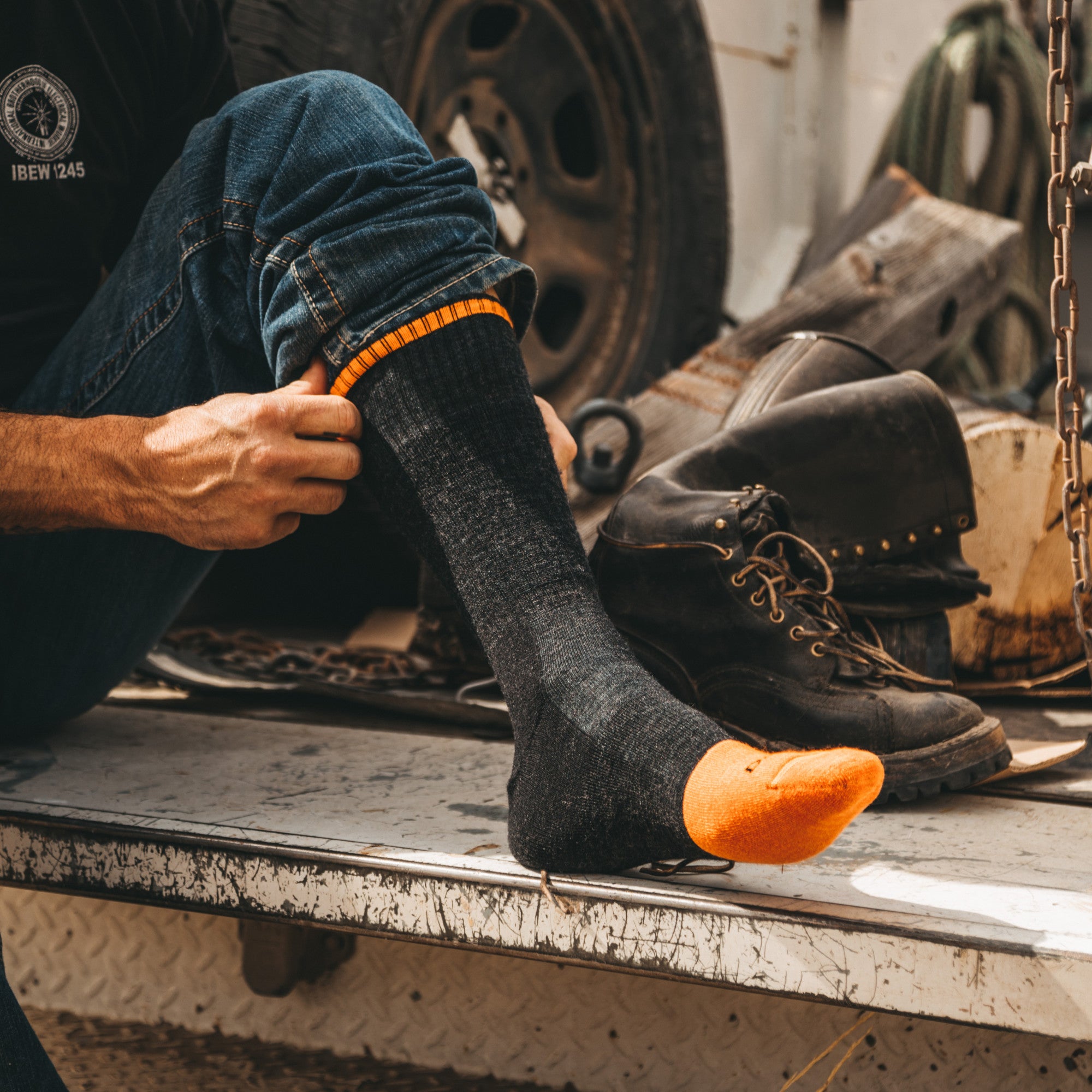 Side shot of model sitting on the back of a truck bed putting on the men's steely boot work socks in graphite