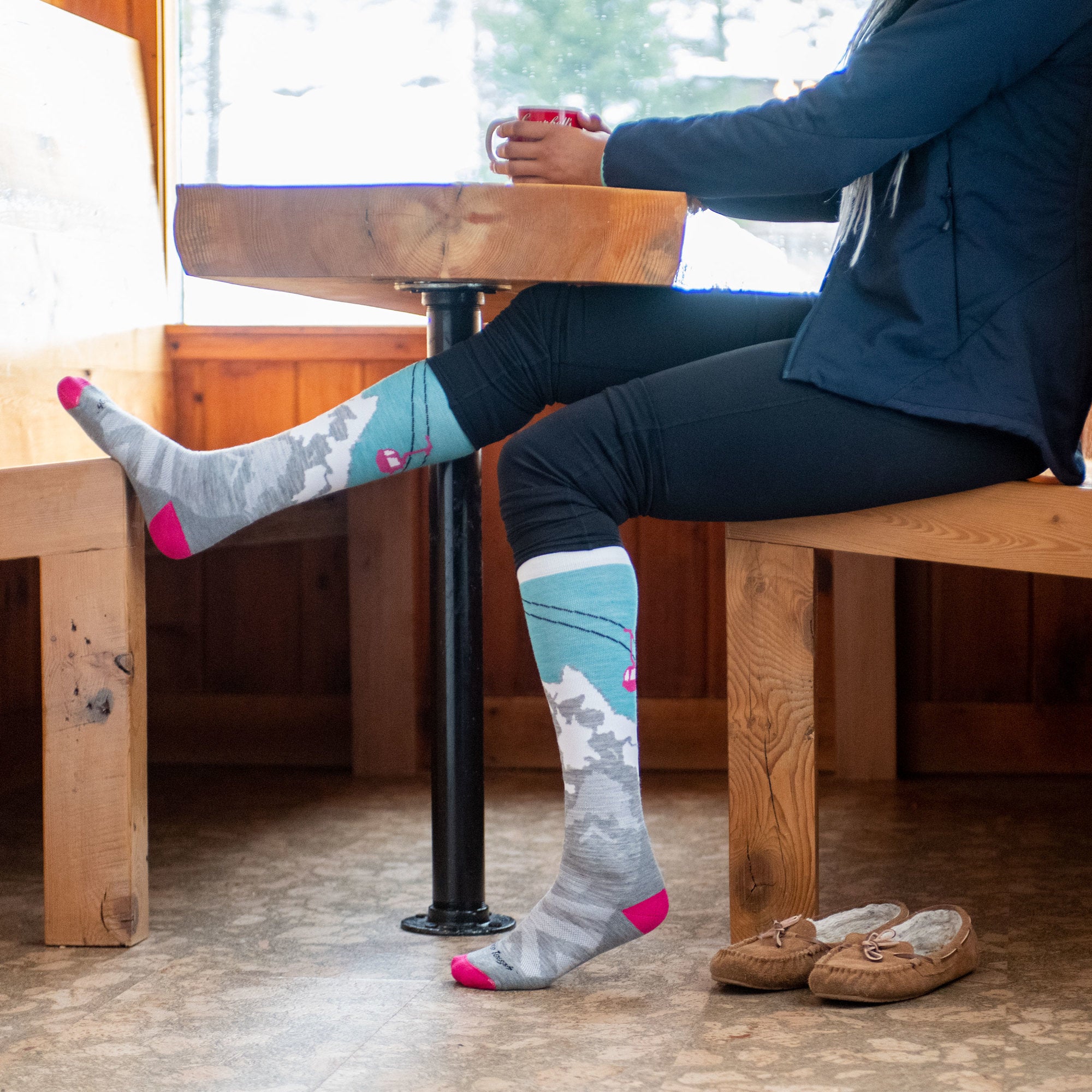 Side shot of model sitting down inside having coffee wearing women's yeti over-the-calf ski sock in aqua