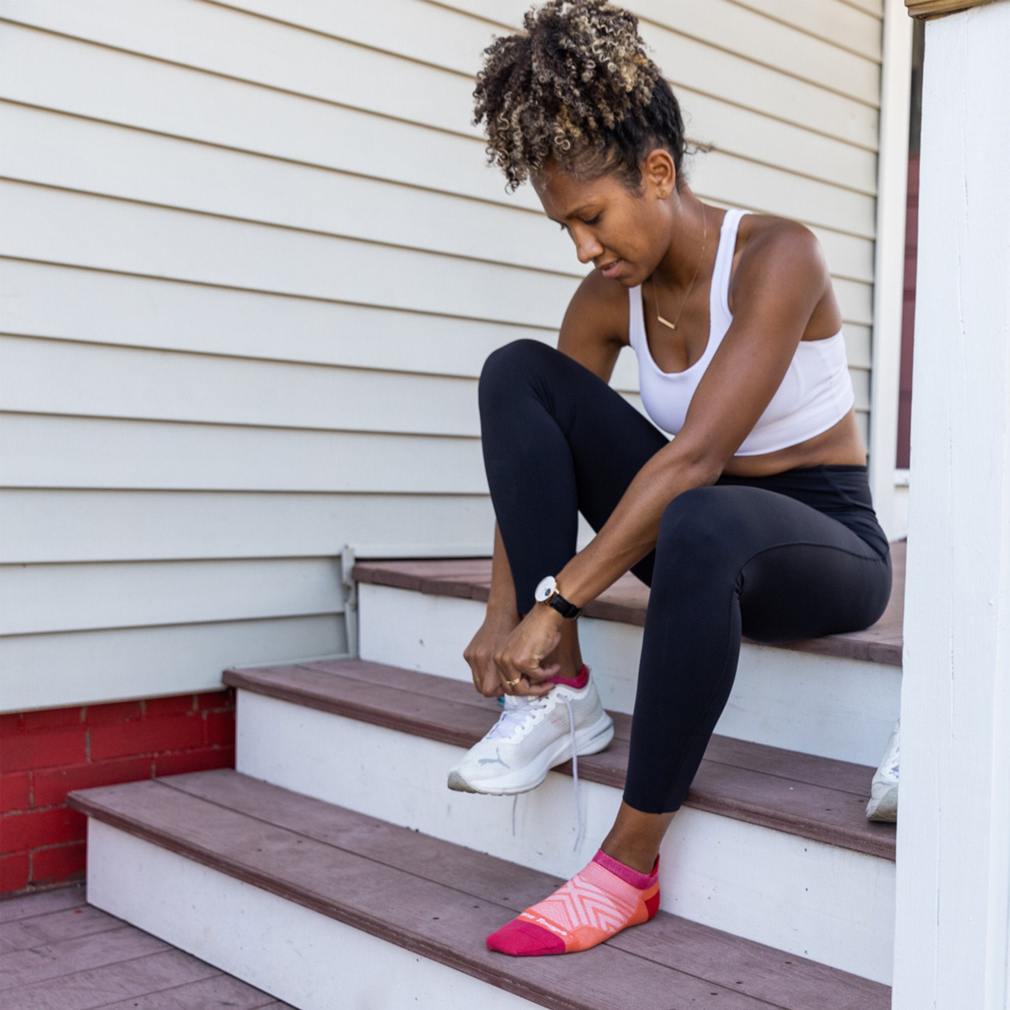 Model sitting on steps putting on her sneakers wearing the women's no show tab running sock in coral pink