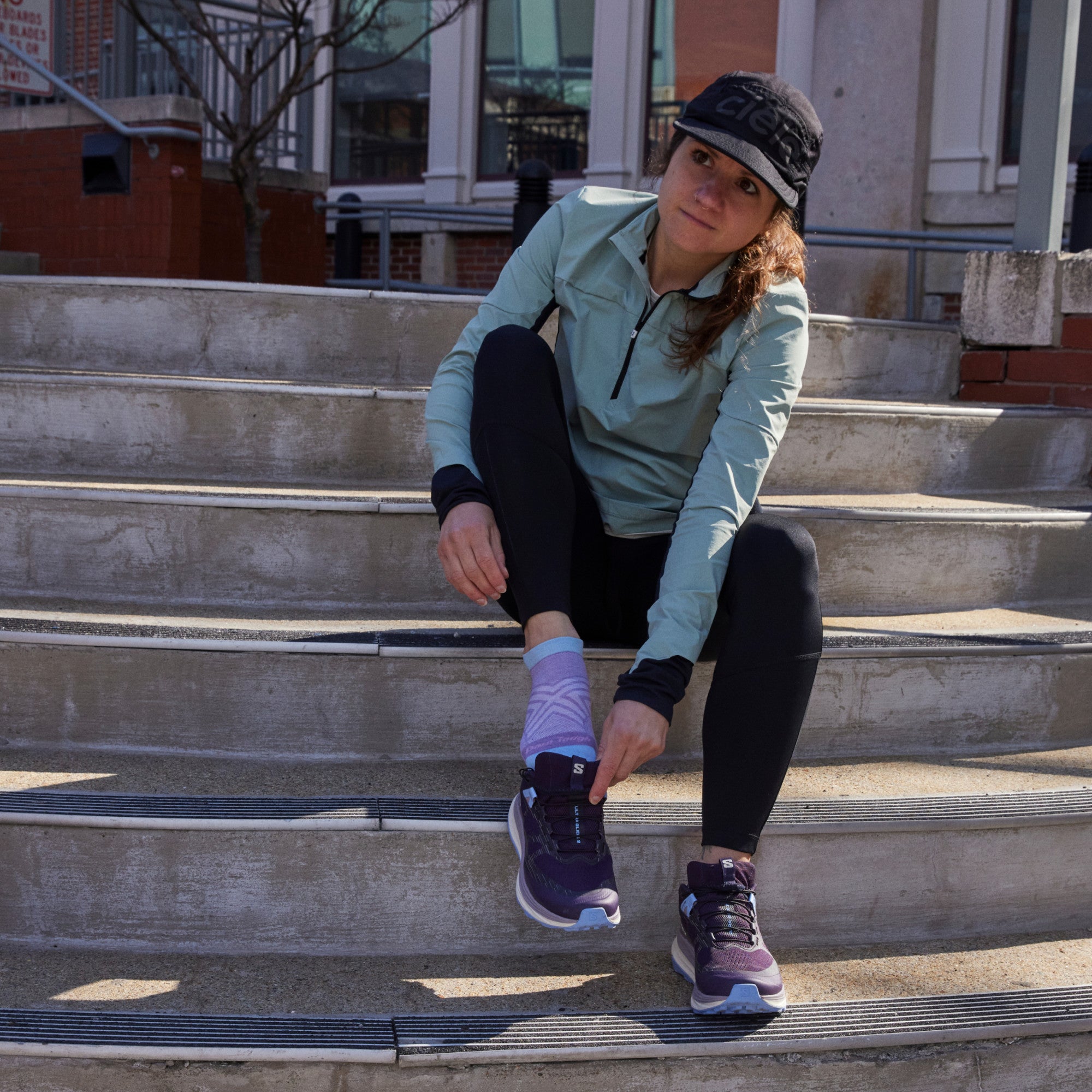 Model sitting on concrete steps while wearing 1043 socks in lavender colorway and slipping on purple running shoe on right foot