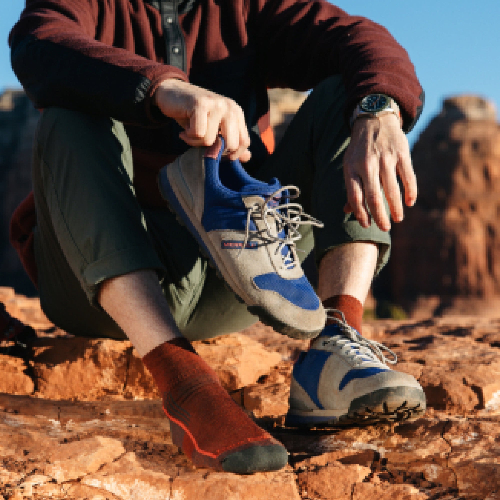 Image of model sitting on red desert rocks wearing 1959 socks in chestnut colorway while wearing one blue hiking shoe and holding the other