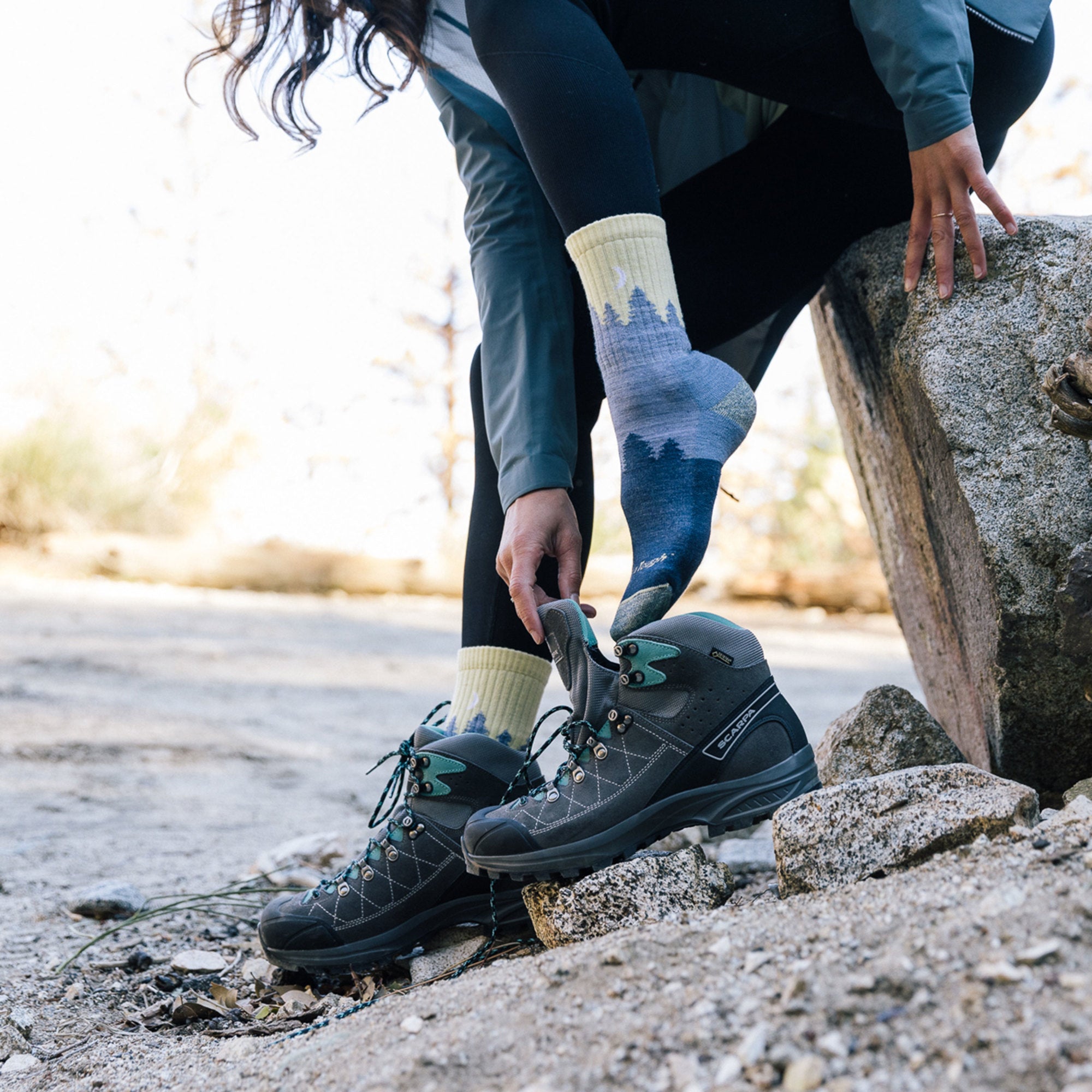 Model sitting on a rock wearing women's treeline micro crew hiking sock in denim while putting on hiking shoes