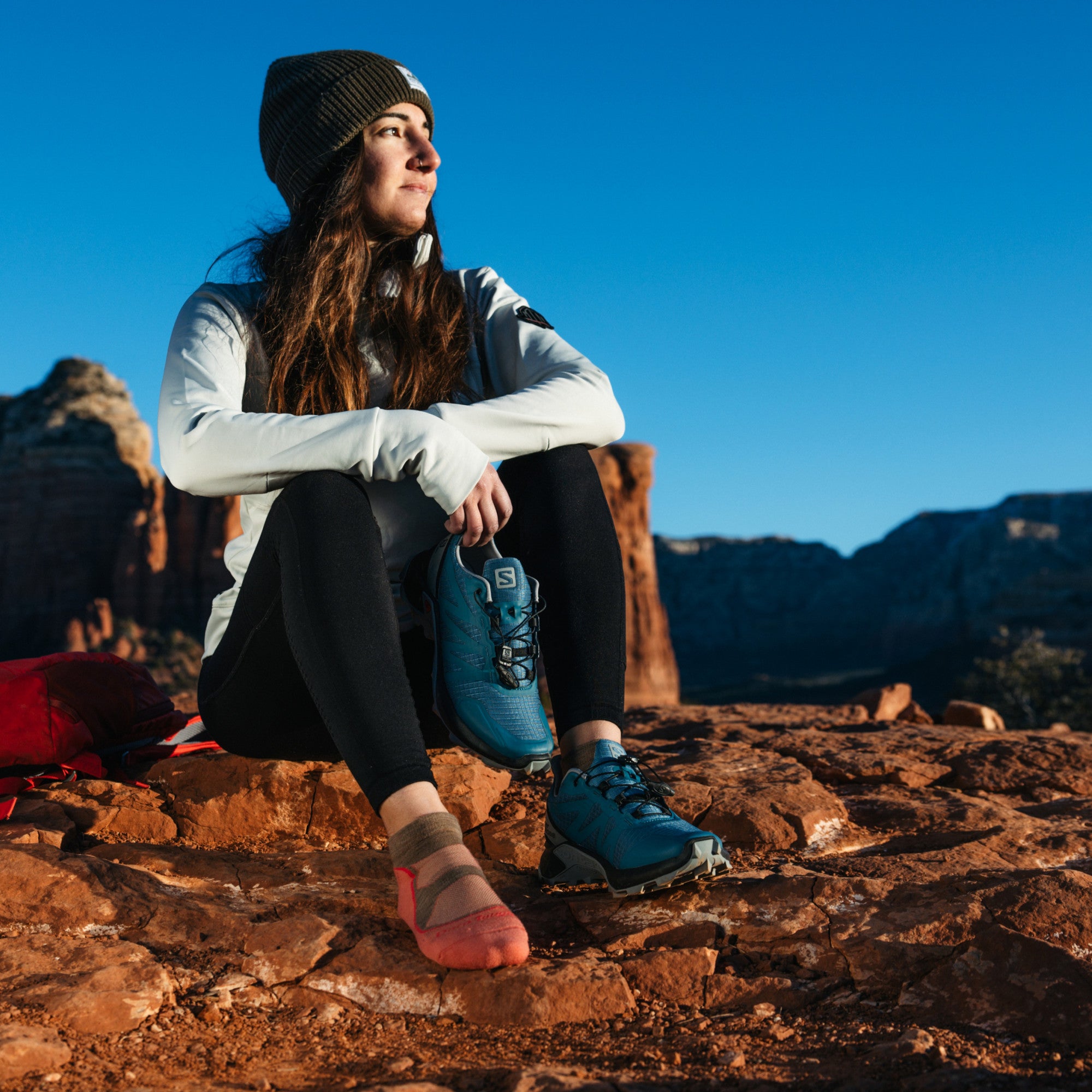 Image of model sitting on red desert rocks wearing 1986 socks in Canyon colorway while wearing one blue hiking shoe and holding the other