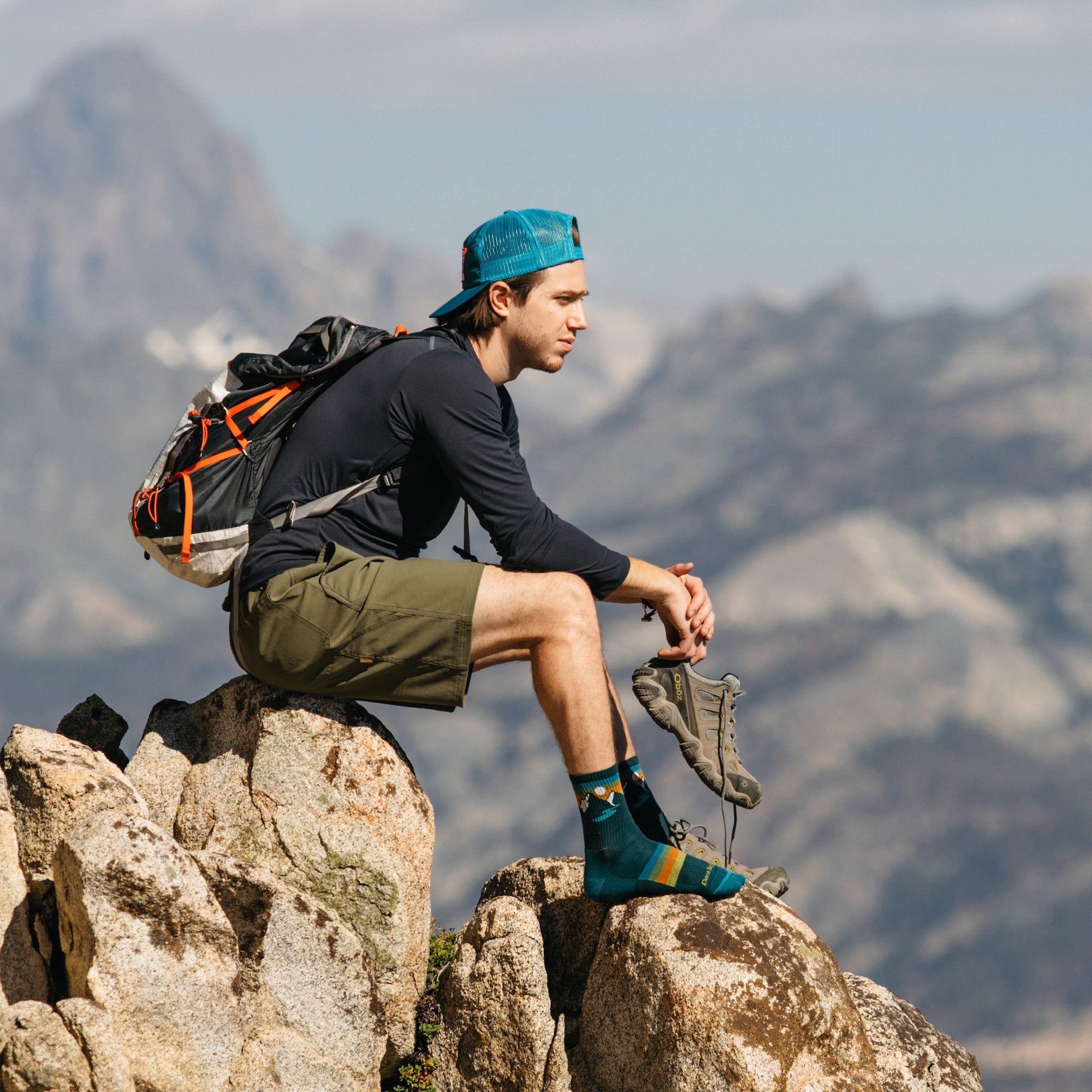 Model sitting on top of a mountain wearing the men's sunset ridge micro crew hiking sock in bottle blue