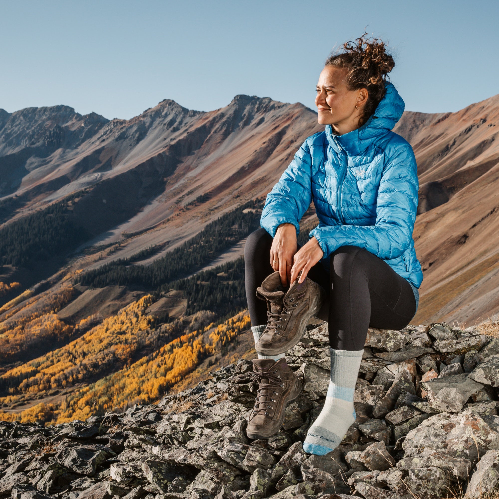 model sitting on mountain rocks holding one shoe in her hands wearing the 5011 micro crew mountaineering sock in reed