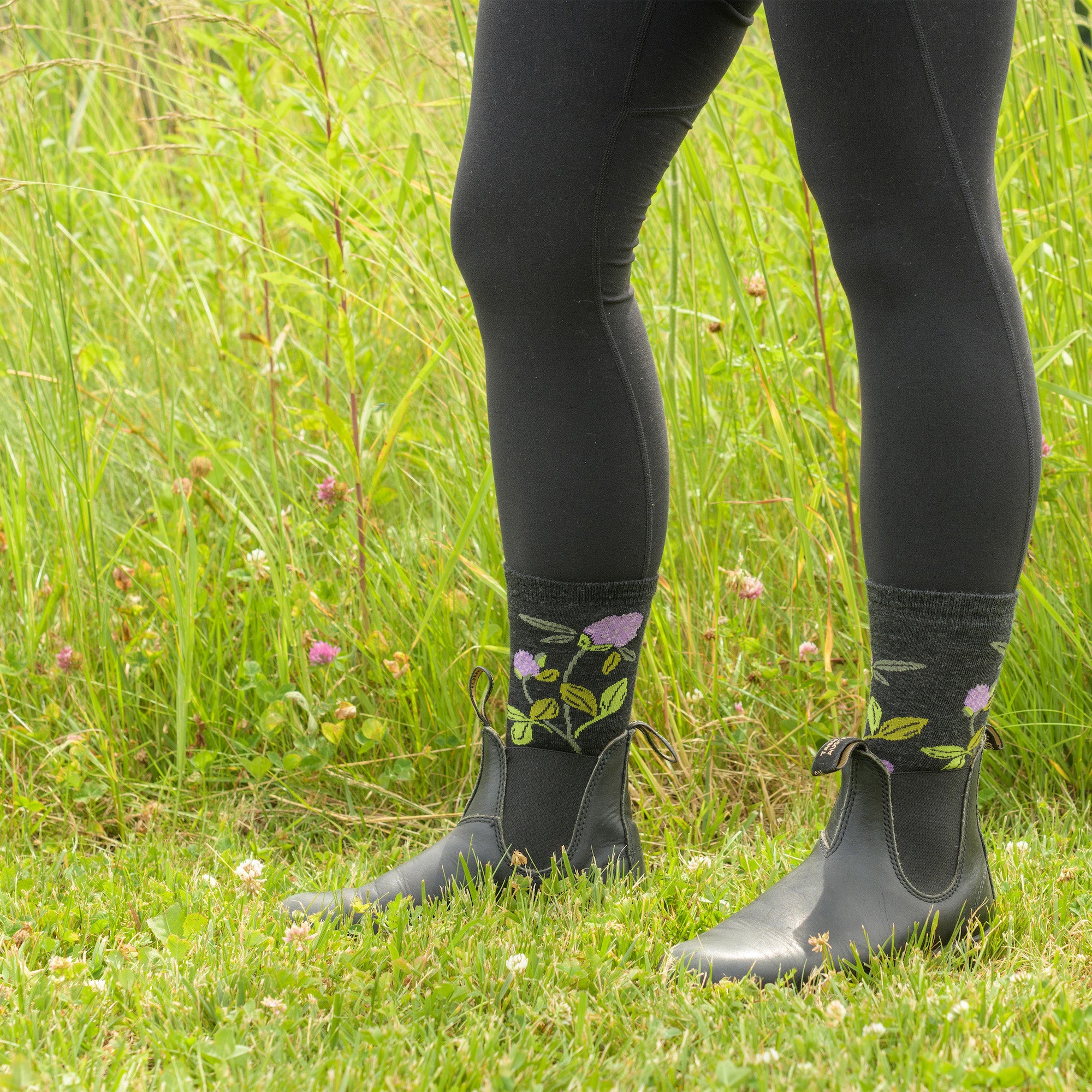 Close up shot of model standing in the grass in rainboots wearing the women's blossom crew lifestyle socks in charcoal