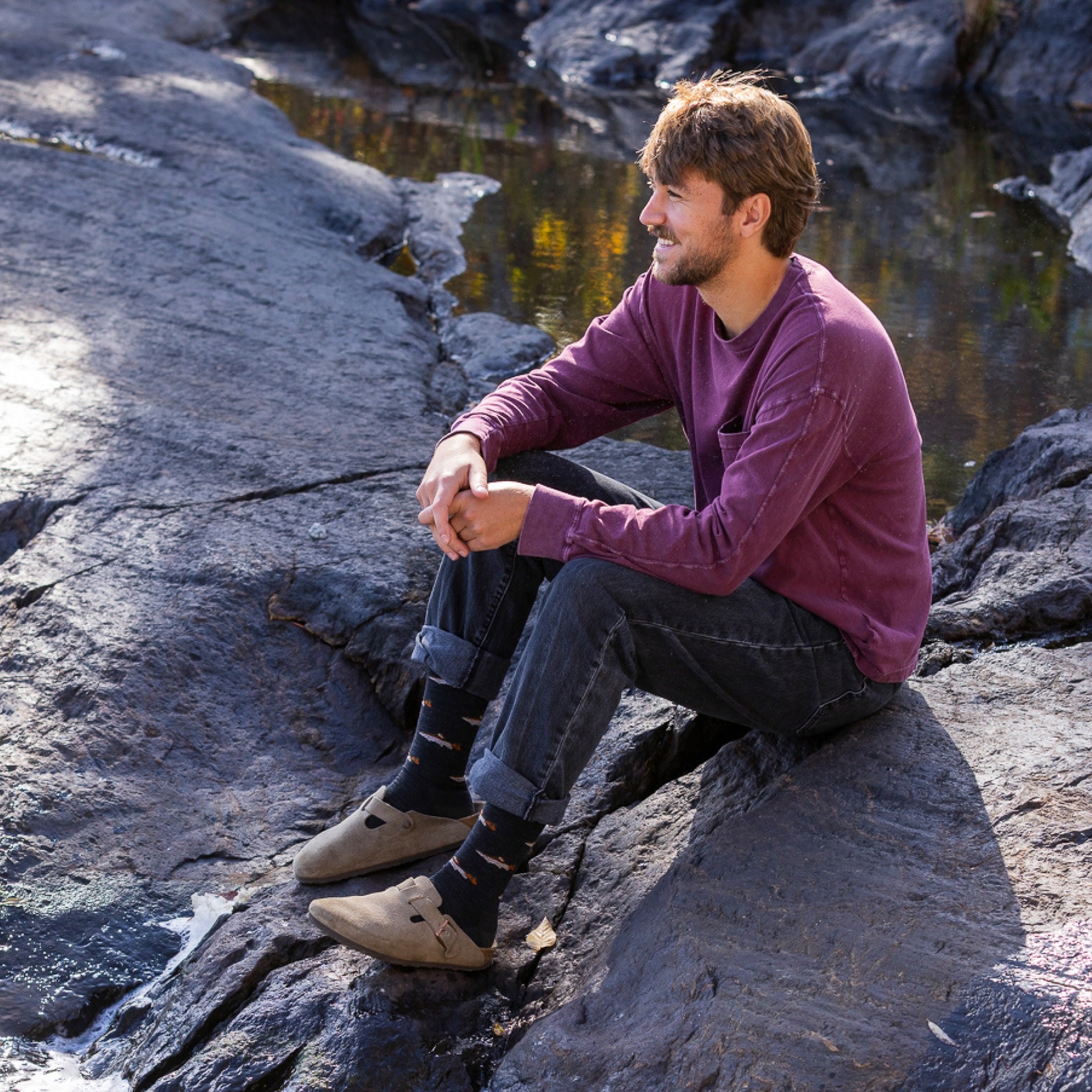 model sitting on rocks next to water wearing the 6085 spey fly lightweight lifestyle crew in charcoal colorway
