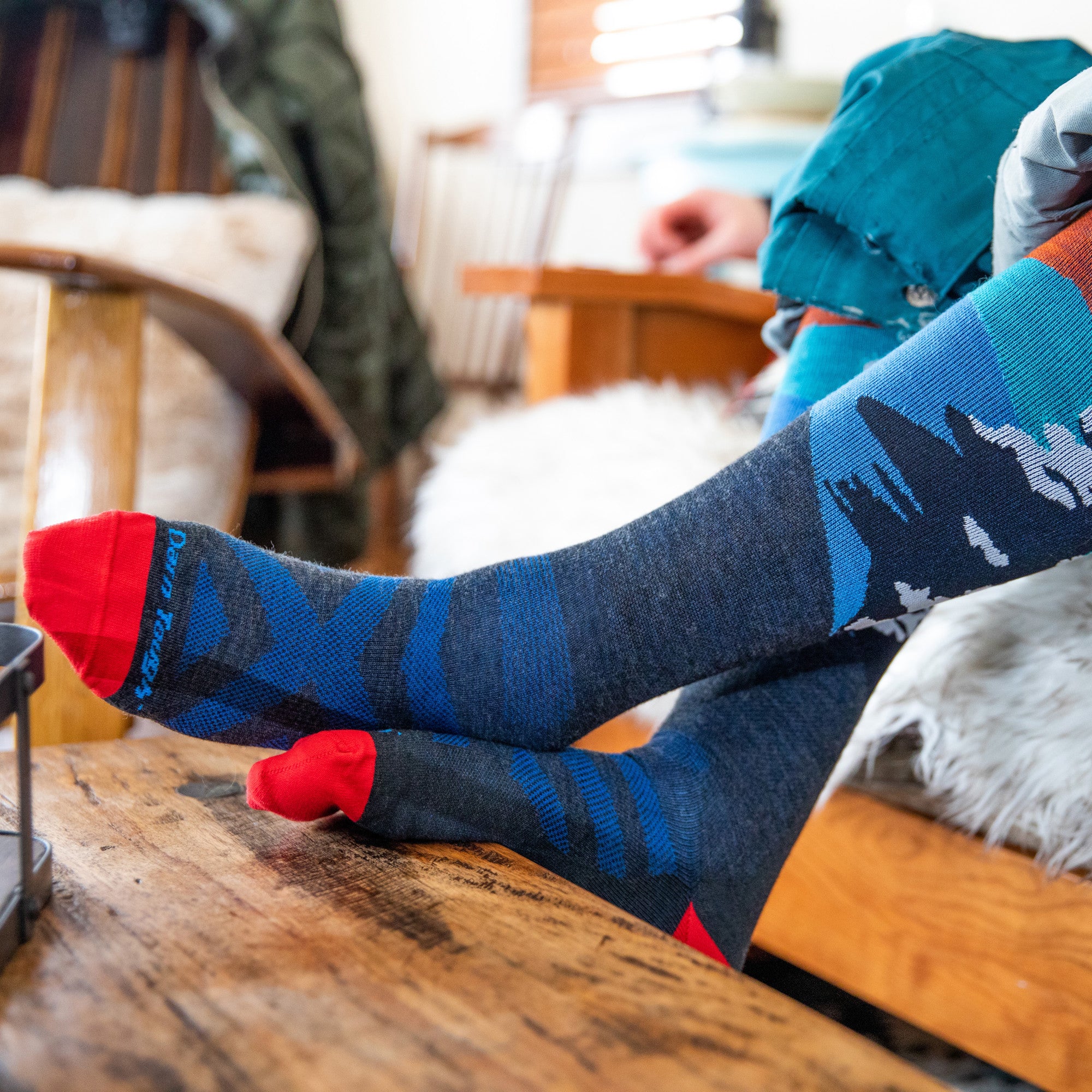 Close up shot of model with their feet up on a coffee table wearing the men's solstice over-the-calf snow sock in charcoal