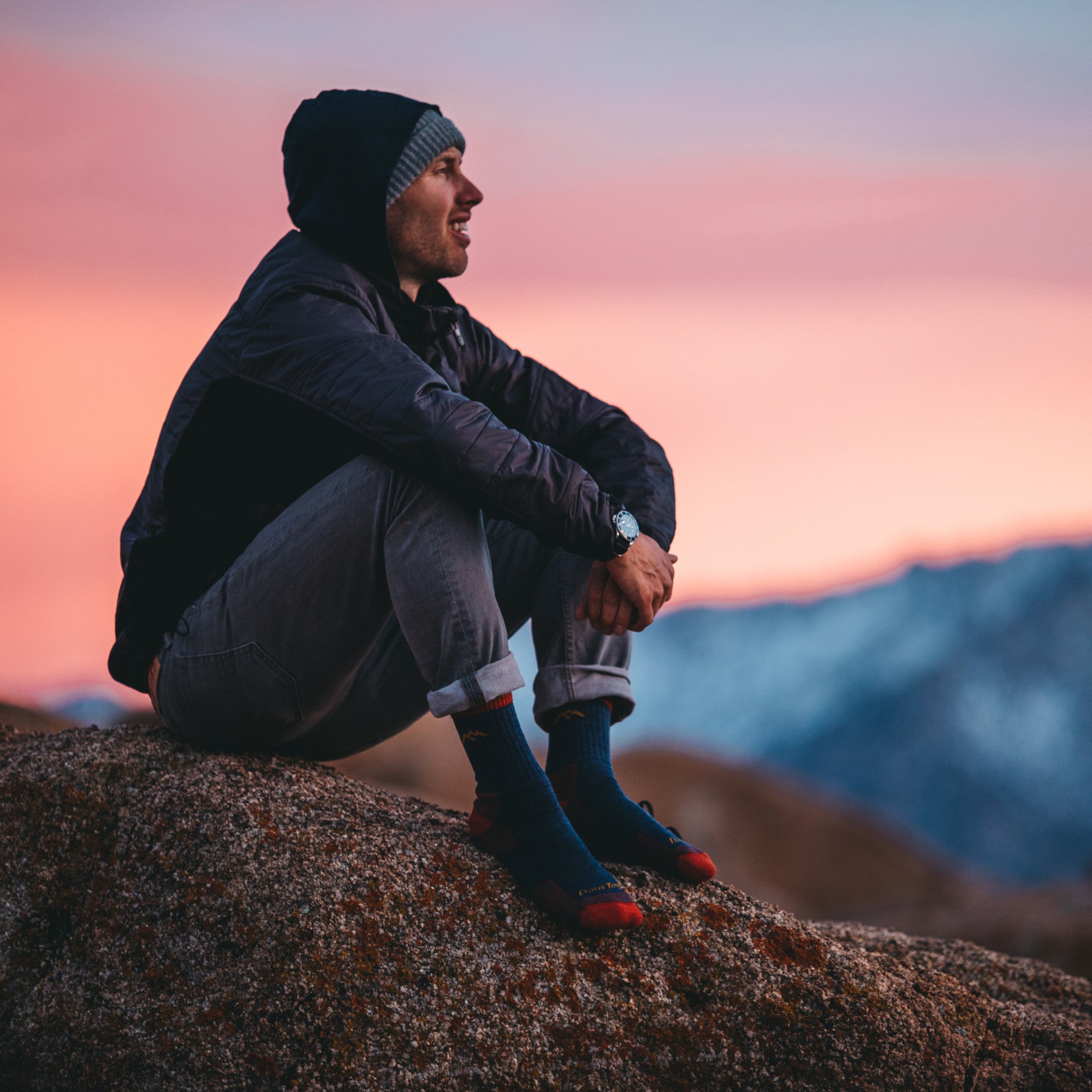Full shot of model sitting on edge of a mountain watching the sunset wearing the men's micro crew hiking sock in denim
