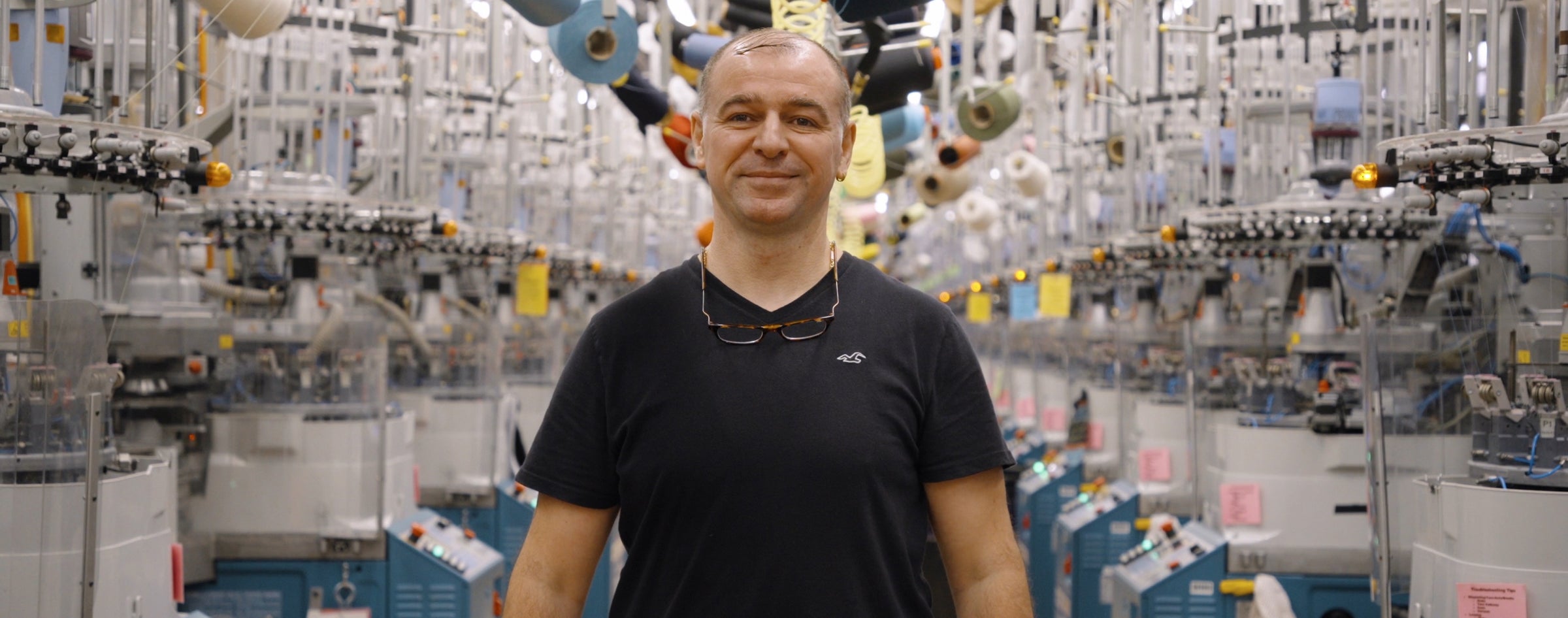 Darn Tough Master Technician Adnan standing on the Mill flour with knitting machines surrounding him