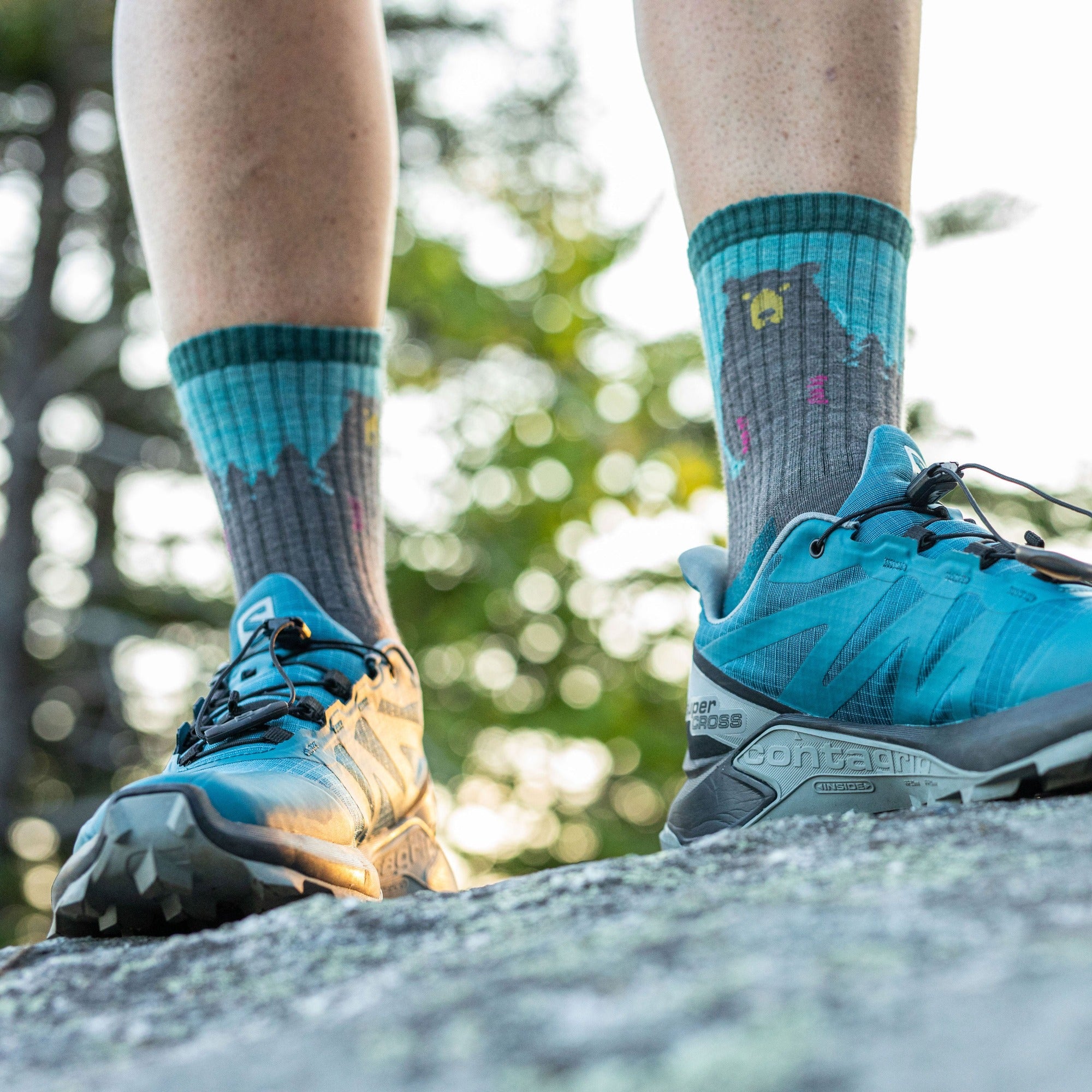 Close up image of a woman's feet, standing on a rock wearing blue sneakers and the women's bear town micro crew hiking sock in aqua